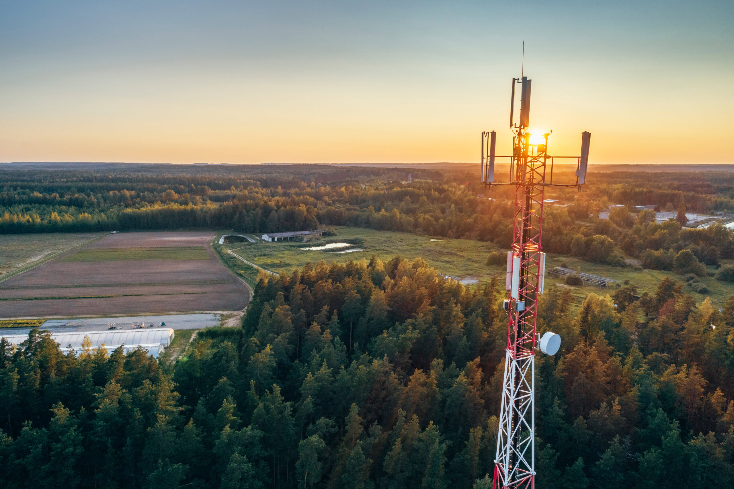 A red and white telecommunications tower designed using OpenTower Designer stands in a forested area at sunset, with fields and more trees in the background.