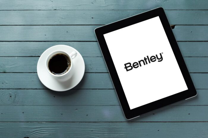 A cup of coffee in a white saucer is placed next to a tablet displaying the Bentley logo on a gray wooden surface.