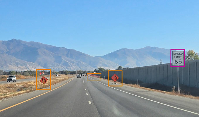 Highway with two lanes, speed limit sign reading 65, and two road work signs on side of the road. Mountains and clear sky in the background.